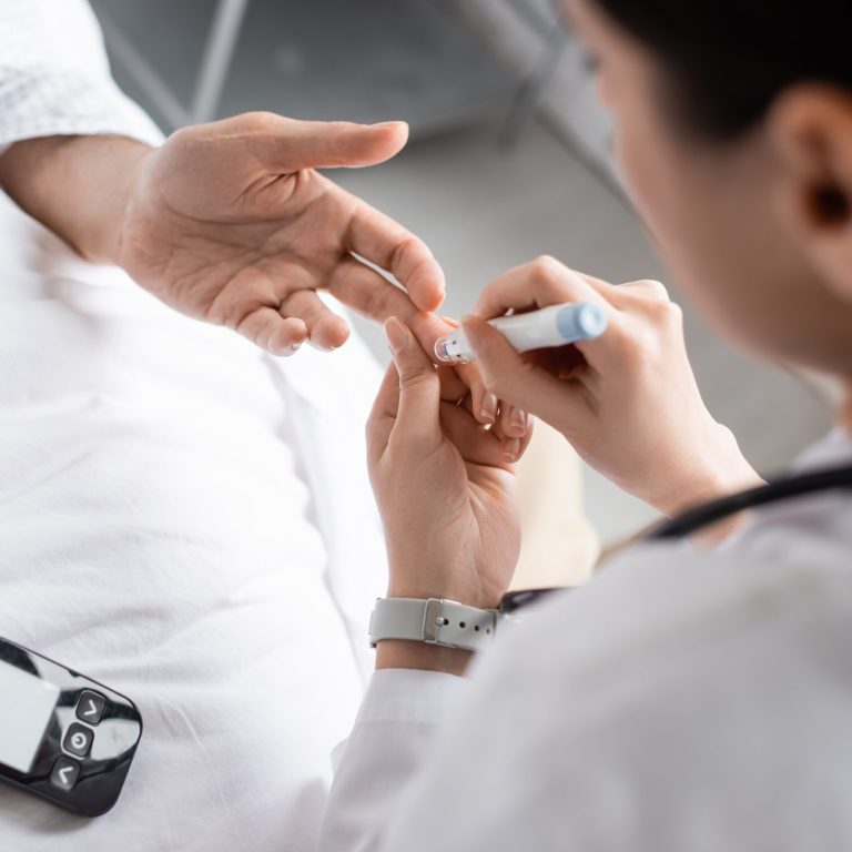 Blurred doctor holding lancet pen near hand of patient with diabetes in hospital ward